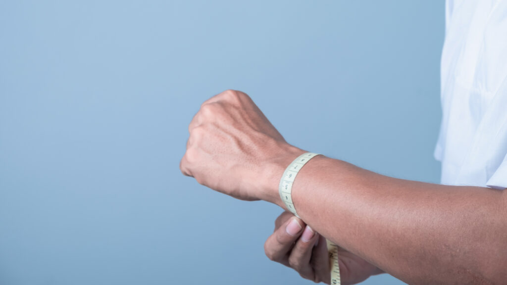 A man measures his wrist with a tape measure on light blue background, weight loss concept